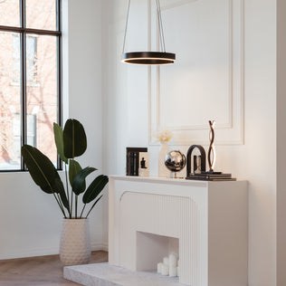 A modern living room features a white mantel adorned with books, a globe, and sculptures. Nearby stands a large potted plant, while the Artika Vance modern LED pendant light 3 CCT hangs above the mantel from the ceiling.