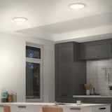 Modern kitchen with gray cabinets, a white tiled backsplash, recessed ceiling lights, a large window, and a white countertop with cups and jars. Natural light is visible outside the window.