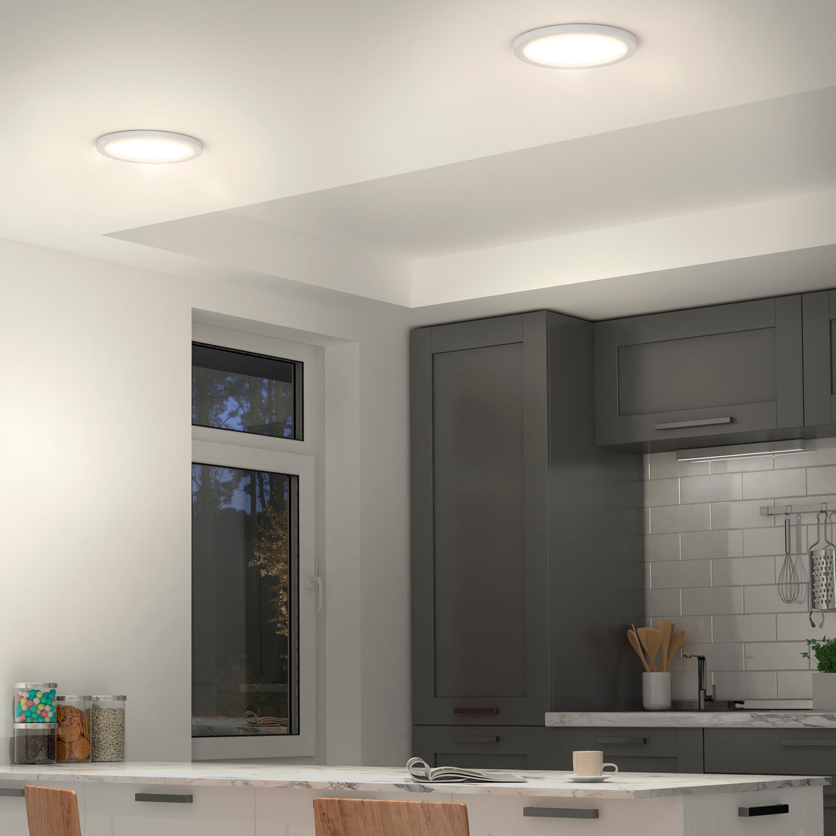 Modern kitchen with gray cabinets, white tile backsplash, and a window with a view of trees. Two Artika Europa Disk 9 in. LED modern flush mount lights brighten the space. A white island with chairs and kitchenware is in front.
