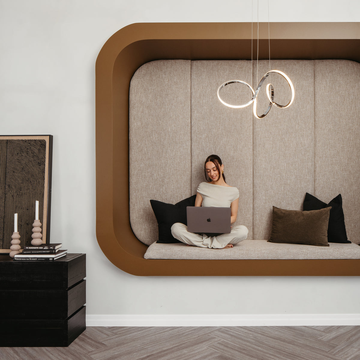 A woman sits cross-legged on a built-in cushioned nook with pillows, using a laptop. Modern pendant lights hang above her, and a side table with candles and artwork is nearby. The decor is minimalist and contemporary.