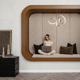 A woman sits cross-legged on a built-in cushioned nook with pillows, using a laptop. Modern pendant lights hang above her, and a side table with candles and artwork is nearby. The decor is minimalist and contemporary.