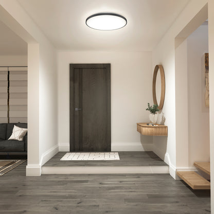 A modern entryway features a dark wooden door, light walls, wood flooring, a round mirror above a floating wooden shelf with a plant, and an Artika Europa 21 in. LED modern flush mount light with remote. The living room is partially visible on the left.