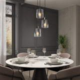 A modern dining room with a round white marble table set for four, surrounded by beige chairs. Three pendant lights hang above the table, and a tall plant stands in the corner near large windows with sheer curtains.