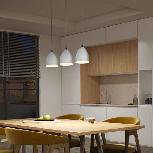 A modern kitchen with wooden accents features the Artika Bergen 3-light LED pendant light above a wooden dining table with yellow chairs, a fruit bowl, and a mug. The kitchen sink is visible in the background.