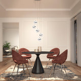 A modern dining room with a round black table, four reddish-brown velvet chairs, a patterned rug, and five glass pendant lights above. Light streams in through sheer curtains. A cozy sitting area is visible in the background.