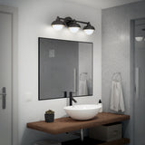 A modern bathroom with a round white vessel sink on a wooden countertop, a black faucet, a small potted plant, a towel ring with a white towel, and a rectangular mirror under a three-bulb light fixture.