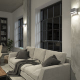A modern living room with a light gray sofa, dark cushions, a throw blanket, large windows, concrete walls, wall-mounted lights, a bookshelf, and a potted plant in the corner.