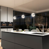 Modern kitchen with a large island, built-in ovens, and three spherical pendant lights. Floor-to-ceiling windows reveal trees outside, and a dining area with chairs is visible in the background.