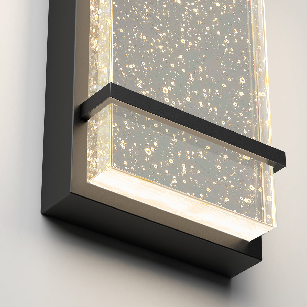Close-up of a modern wall sconce featuring a rectangular glass panel with tiny bubbles inside, illuminated by soft white lights, and framed with sleek black metal.