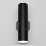 A modern black cylindrical wall sconce with small star-like holes and a glowing white light at the bottom, mounted on a plain light gray wall.
