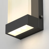A close-up of a modern wall-mounted light fixture with a rectangular black frame and a frosted cover, emitting a soft white glow from three LED bulbs.