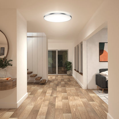 A modern hallway features wooden floors, light walls, Artika’s Europa 21 in. LED flush mount with remote, a round mirror above a console table with a plant, glass-railed stairs, and a view of the living room with couch and art.