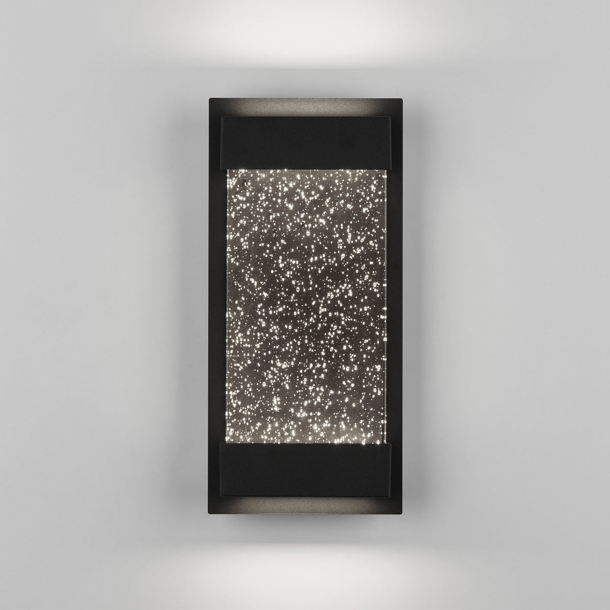 The Artika Essence Glow Box indoor-outdoor LED wall light features a modern black frame and rectangular design, casting up-and-down lighting through a textured panel filled with star-like dots for a sparkling effect on gray walls.