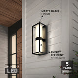 A modern outdoor wall light with a matte black finish and integrated energy-efficient LED, mounted on light wood siding. Text highlights features and a 5-year warranty badge is visible in the corner.