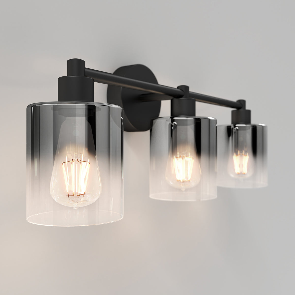 A modern wall-mounted light fixture with three exposed Edison bulbs, each enclosed in a clear cylindrical glass shade, is attached to a matte black metal frame against a light gray wall.