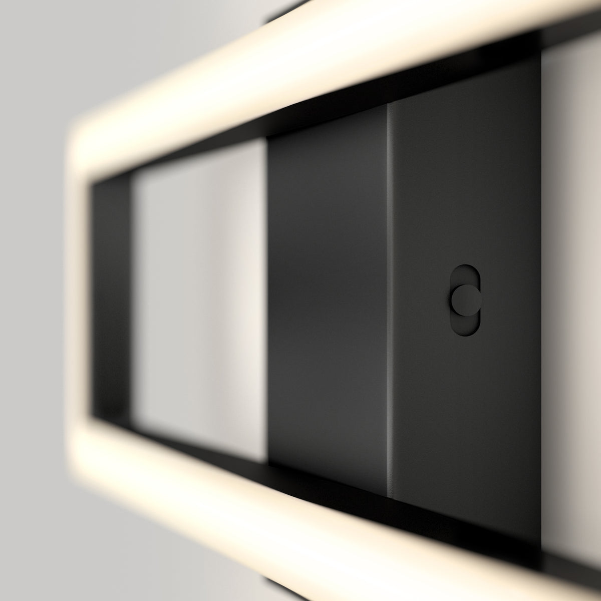 A close-up view of a modern, rectangular black light fixture with soft white LED lighting along its edges, mounted on a light gray wall. The design is minimalistic and geometric.