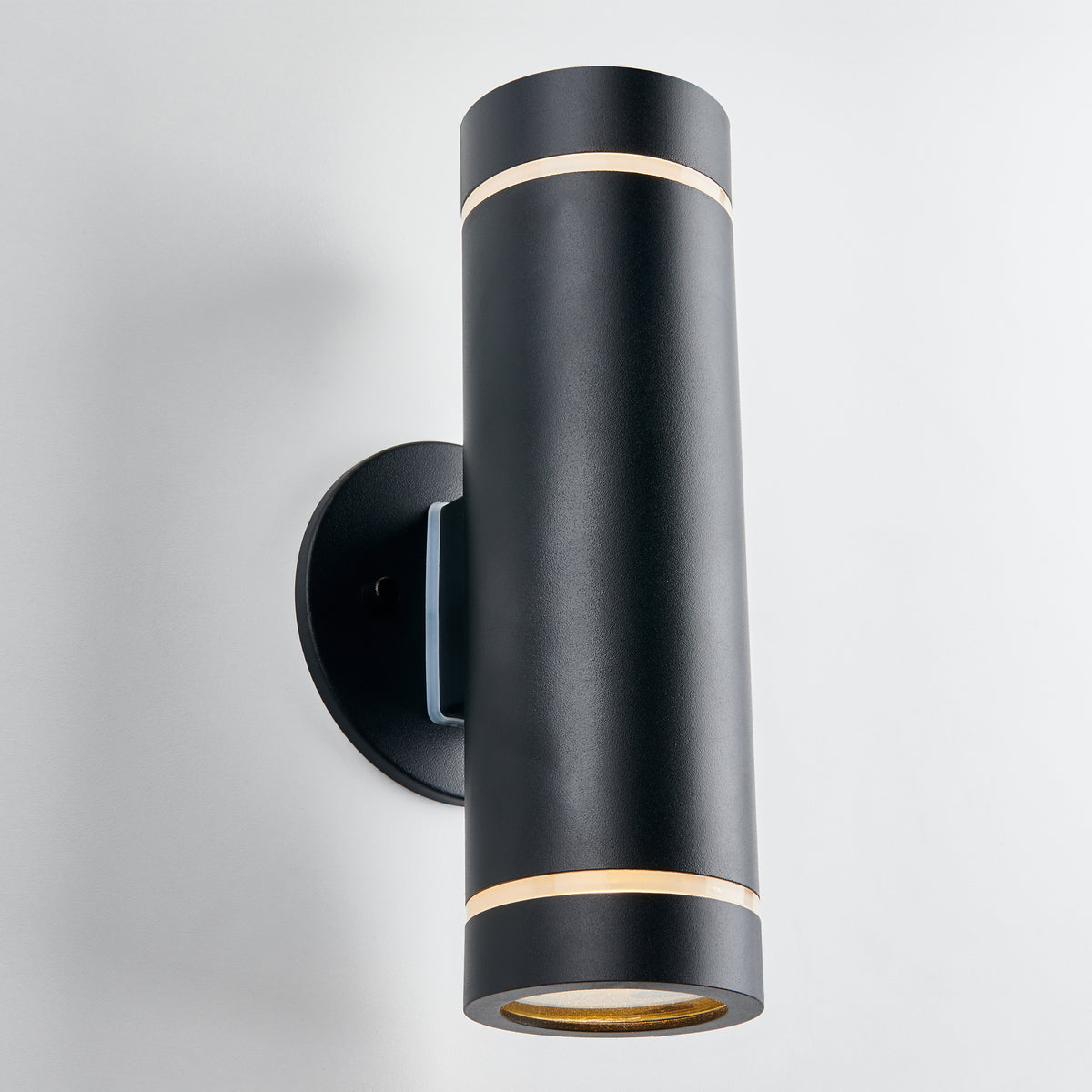 The Artika C7 indoor-outdoor wall light features a modern cylindrical black design with a matte finish and slim gold bands at each end, mounted on a round base against a white background.