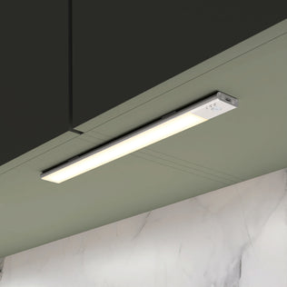 The Artika Outline under cabinet LED light 3CCT, a slim rectangular fixture, is mounted beneath a dark kitchen cabinet, brightening the light green wall and white marble backsplash below.