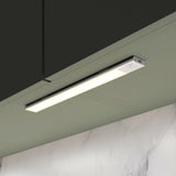 A slim, rectangular LED light fixture is mounted under a dark green cabinet, illuminating a light green wall and a marble backsplash below.