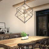 A modern dining room with a rustic wooden table, black cross-back chairs, a geometric metal pendant light, potted plants as a centerpiece, framed art on a sideboard, and large windows letting in natural light.