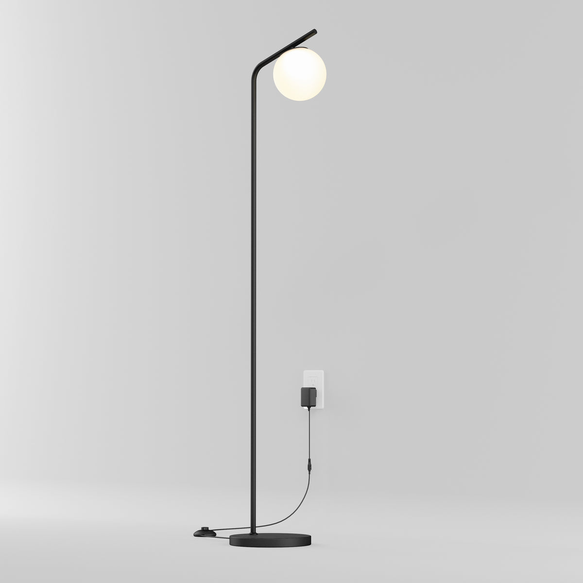 A modern, minimalist black floor lamp with a round white bulb at the top stands near a white wall outlet. The lamp is plugged in, and the cord is visible running to the socket. The background is plain and light-colored.