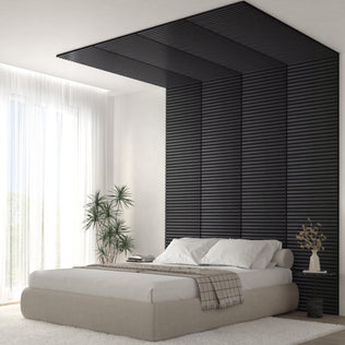 A modern bedroom features a beige bed, white bedding, and the Artika Sonolok 15.5 ft sound-absorbing wall panel kit as a black slatted accent wall extending onto the ceiling, with large windows and potted plants bringing in light and greenery.