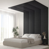 A modern bedroom with a low beige bed, white bedding, and a textured black accent wall that extends onto the ceiling. A potted plant and a vase with branches sit nearby; sheer curtains allow natural light to fill the room.