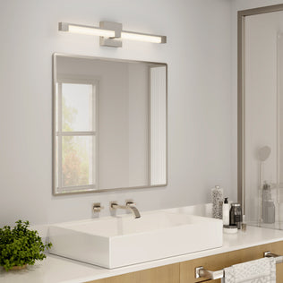 A modern bathroom features a square white sink, wall-mounted faucet, framed mirror, and the Artika Tivoli modern LED bathroom vanity light 5 CCT above the mirror. Toiletries, towel, small green plant on the counter. Natural light comes through a window.