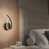 A modern bedroom features a tufted headboard, a wall-mounted abstract light fixture, a neatly made bed with gray bedding and pillows, and a bedside table holding a book and a coffee cup.