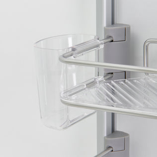 Close-up of an Artika O2 hanging Shower Caddy featuring a transparent plastic cup on a silver shelf with metallic rails, mounted to a vertical grey pole against a light background.