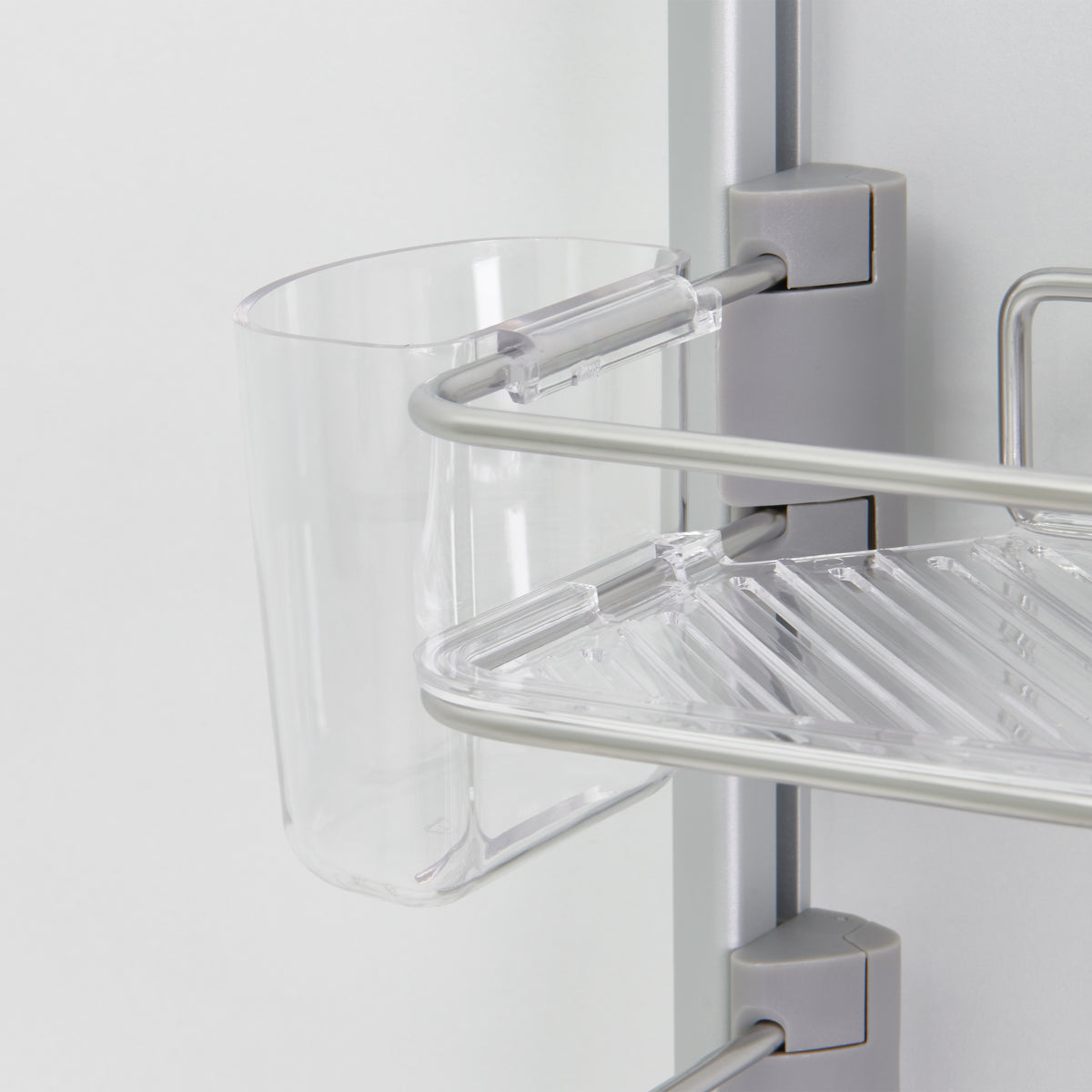 A close-up of a transparent plastic cup attached to a silver and clear plastic bathroom shelf unit, mounted on a light-colored wall. The shelf has a slatted design for drainage.