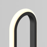 A close-up of a modern, minimalist lamp with a black frame and a glowing, curved white light on a plain gray background.