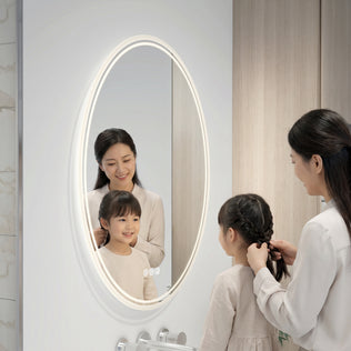 A woman braids a young girl’s hair in front of the Artika Corby 22 in. x 28 in. LED mirror with defogger, featuring adjustable brightness. Their light outfits and reflections are softly illuminated by the mirror’s backlit design.