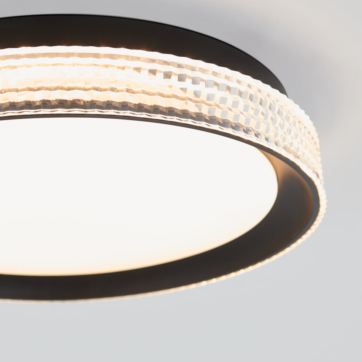 A modern circular ceiling light fixture with a frosted white center and a decorative, textured clear outer ring, emitting a soft, even glow. The fixture has black trim and is mounted on a white ceiling.