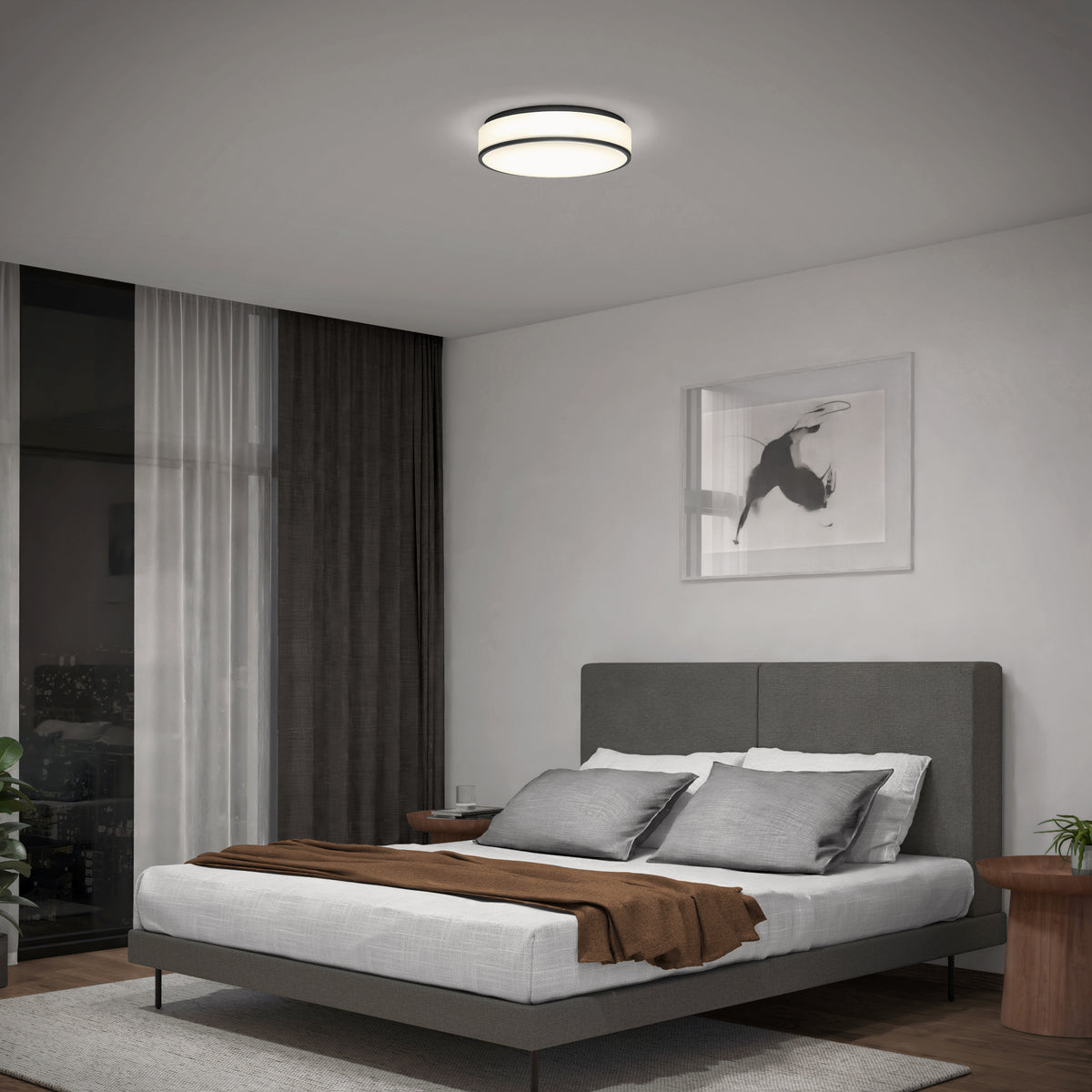 A modern bedroom with a gray upholstered bed, brown throw blanket, and multiple pillows. There is a circular ceiling light, framed artwork above the bed, full-length curtains, and a view of a balcony outside.
