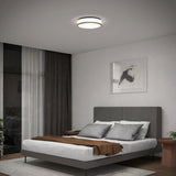 A modern bedroom with a gray upholstered bed, brown throw blanket, and multiple pillows. There is a circular ceiling light, framed artwork above the bed, full-length curtains, and a view of a balcony outside.