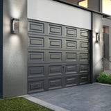 A modern gray garage door with geometric panels is set in a concrete exterior wall, flanked by two rectangular wall lights. There is a small grassy area and steps with a glass railing nearby.