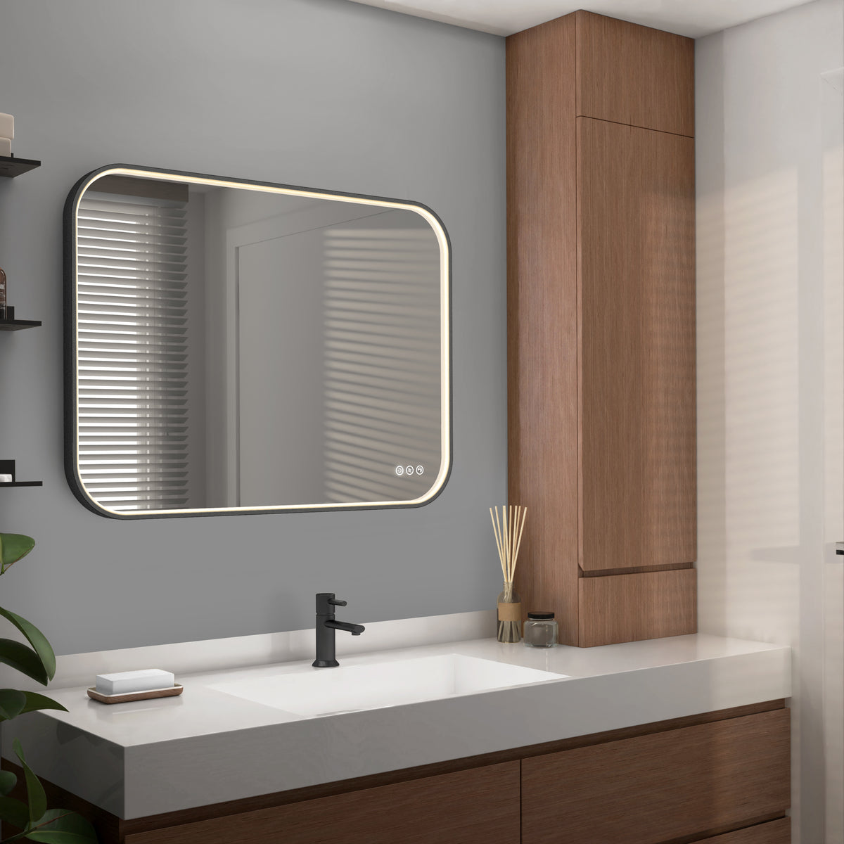 A modern bathroom features the Artika Royce Dimmable LED Mirror (3 CCT, mounts horizontally or vertically) above a white sink with a black faucet, wooden cabinets, a reed diffuser, and a small plant. Light enters through window blinds.