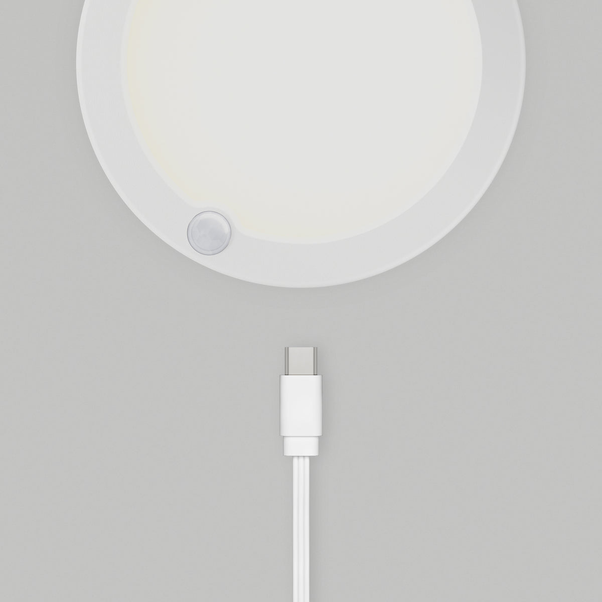 A white USB-C charging cable lies below the Artika Accent under cabinet LED light 3CCT, a round white device with a small circular sensor on its edge, all placed on a light gray surface.