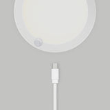 A white USB-C charging cable positioned below a round, white electronic device mounted on a light gray wall. The device has a small circular sensor or button near its edge.