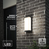 A modern rectangular LED wall light with a matte black finish is mounted on a brick exterior wall. Text highlights its energy efficiency, no bulbs required, and a 3-year warranty. Some greenery is visible below.