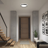 A modern entryway with a wooden front door, minimalist staircase, potted plant, wall art, a cushioned bench, and a round ceiling light fixture. The space features wood flooring and neutral colors.