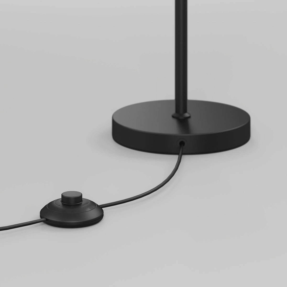 A close-up of a black floor lamp base with a round, black foot switch connected by a cord, set against a plain light gray background.