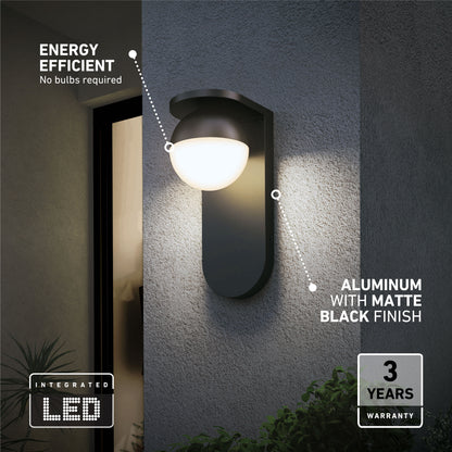 The Artika Bloom indoor-outdoor LED wall light 3 CCT, featuring a matte black aluminum finish, is mounted beside a door. Text notes its energy efficiency, integrated LED, and 3-year warranty. Greenery appears at the bottom right.