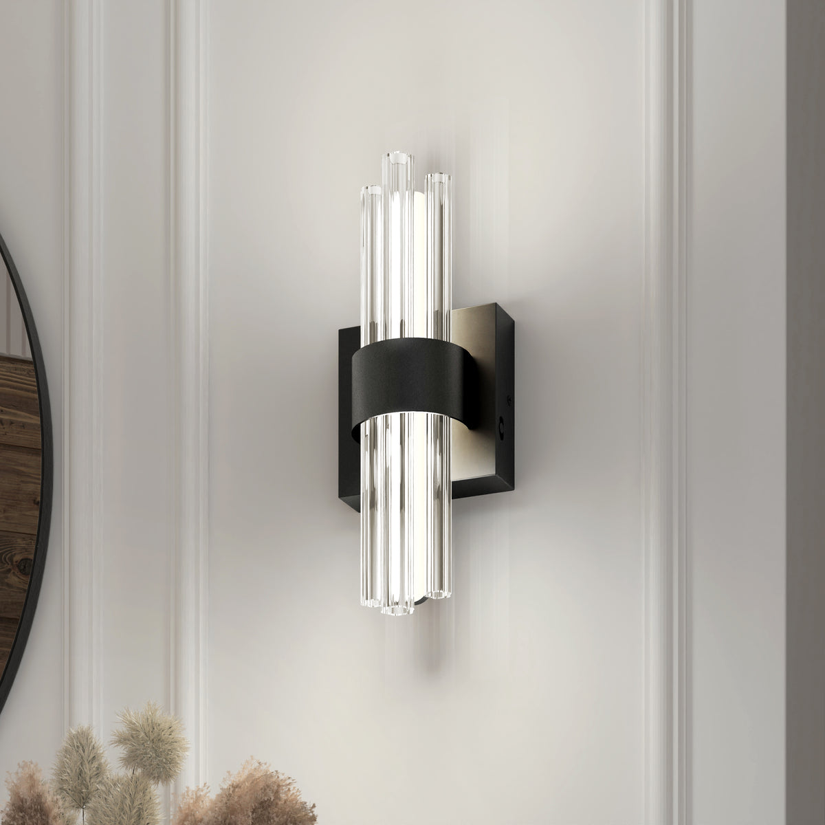 The Artika Dorado contemporary LED wall sconce 3 CCT features vertical clear glass rods and a black metal band on a square base, casting soft light on a white paneled wall with dried plants accenting the bottom left corner.