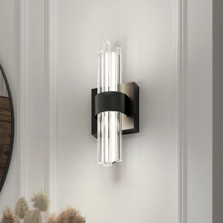 The Artika Dorado contemporary LED wall sconce 3 CCT features vertical clear glass rods and a black metal band on a square base, casting soft light on a white paneled wall with dried plants accenting the bottom left corner.