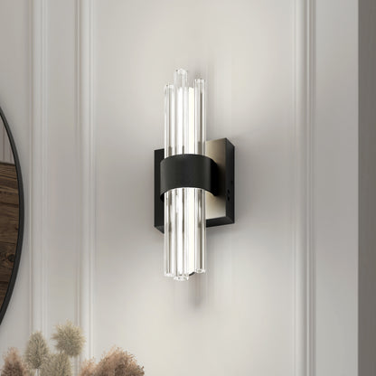 The Artika Dorado LED Contemporary Wall Sconce 3 CCT features vertical glass rods and a black metal band, mounted on a white paneled wall and casting soft, diffused light. Ornamental dried grasses appear in the lower left corner.