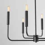 A modern black metal chandelier with four exposed filament light bulbs, each mounted on a straight arm extending from a central cylinder, against a plain light gray background.