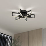 A modern ceiling light fixture with a geometric star-shaped black frame and integrated LED strips, mounted on a white ceiling above wooden cabinets and near a window with black blinds.