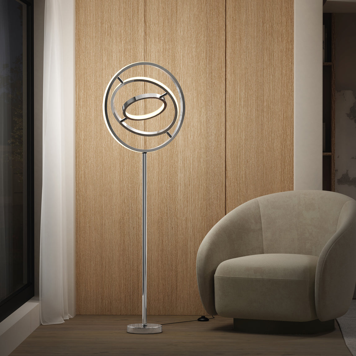 The Artika Pivot modern LED floor lamp 3 CCT, with a metallic circular frame, stands beside a beige curved armchair in a minimalist room featuring wood panel walls, a large window, and sheer white curtains.